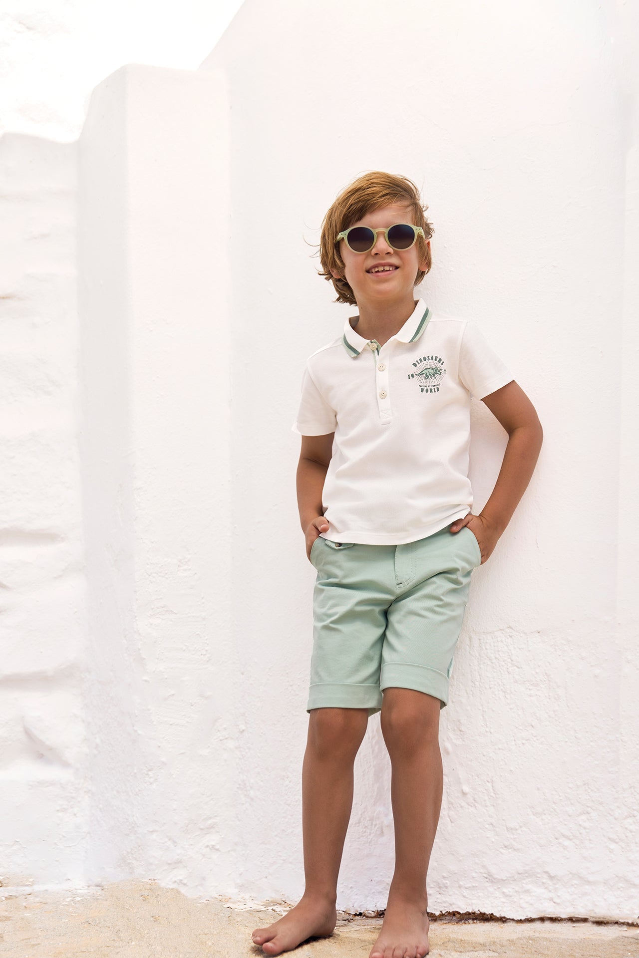A young boy with sunglasses, dressed in a white polo shirt and light green Boys’ Cotton Short from Tartine et Chocolat, stands barefoot against a white wall with his hands in his pockets, smiling. The breathable fabric of the shorts ensures comfort for active play all day long.
