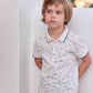 A young child with light brown hair stands against a white wall, wearing the Tartine et Chocolat Boys’ Polo Shirt—a soft cotton white polo featuring a playful dinosaur pattern—paired with green shorts, and gazes off to the side with a neutral expression.