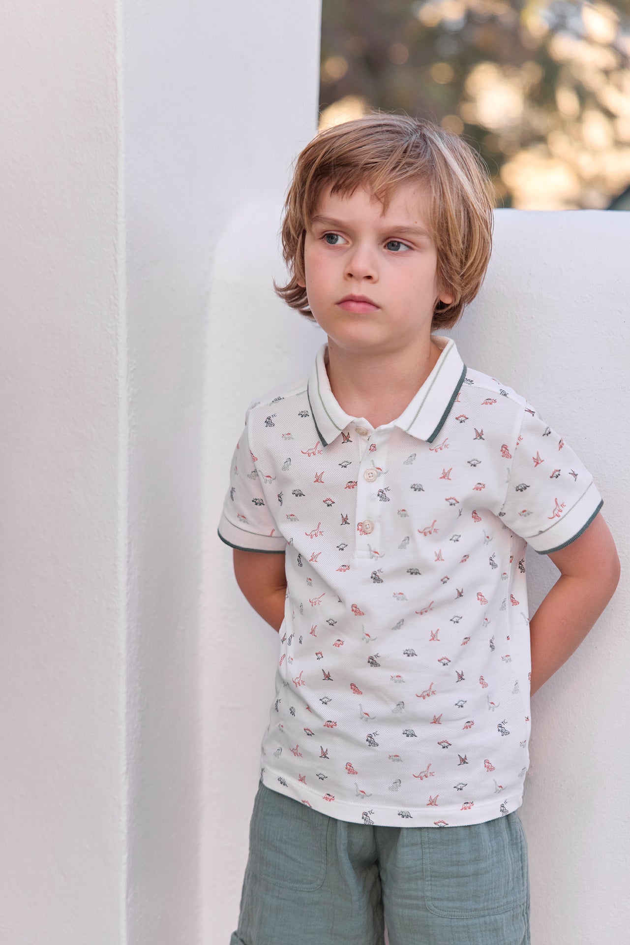 A young child with light brown hair stands against a white wall, wearing the Tartine et Chocolat Boys’ Polo Shirt—a soft cotton white polo featuring a playful dinosaur pattern—paired with green shorts, and gazes off to the side with a neutral expression.
