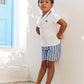 A young boy stands barefoot on white steps, wearing the Tartine et Chocolat Baby Boys’ and Boys’ Polo Shirt and blue striped shorts, with his hands behind his back.