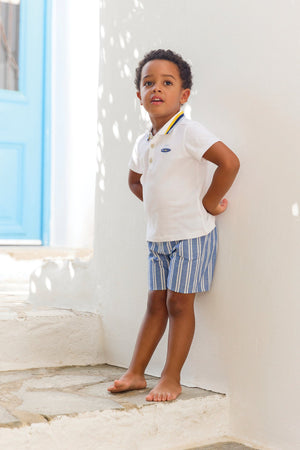 A young boy stands barefoot on white steps, wearing the Tartine et Chocolat Baby Boys’ and Boys’ Polo Shirt and blue striped shorts, with his hands behind his back.