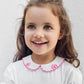 A young child with long brown hair and green eyes smiles while wearing the Tartine et Chocolat Baby Girls’ Peter Pan Collared Onesie, featuring a delicately embroidered white collar with pink flowers, standing against a plain light-colored wall.
