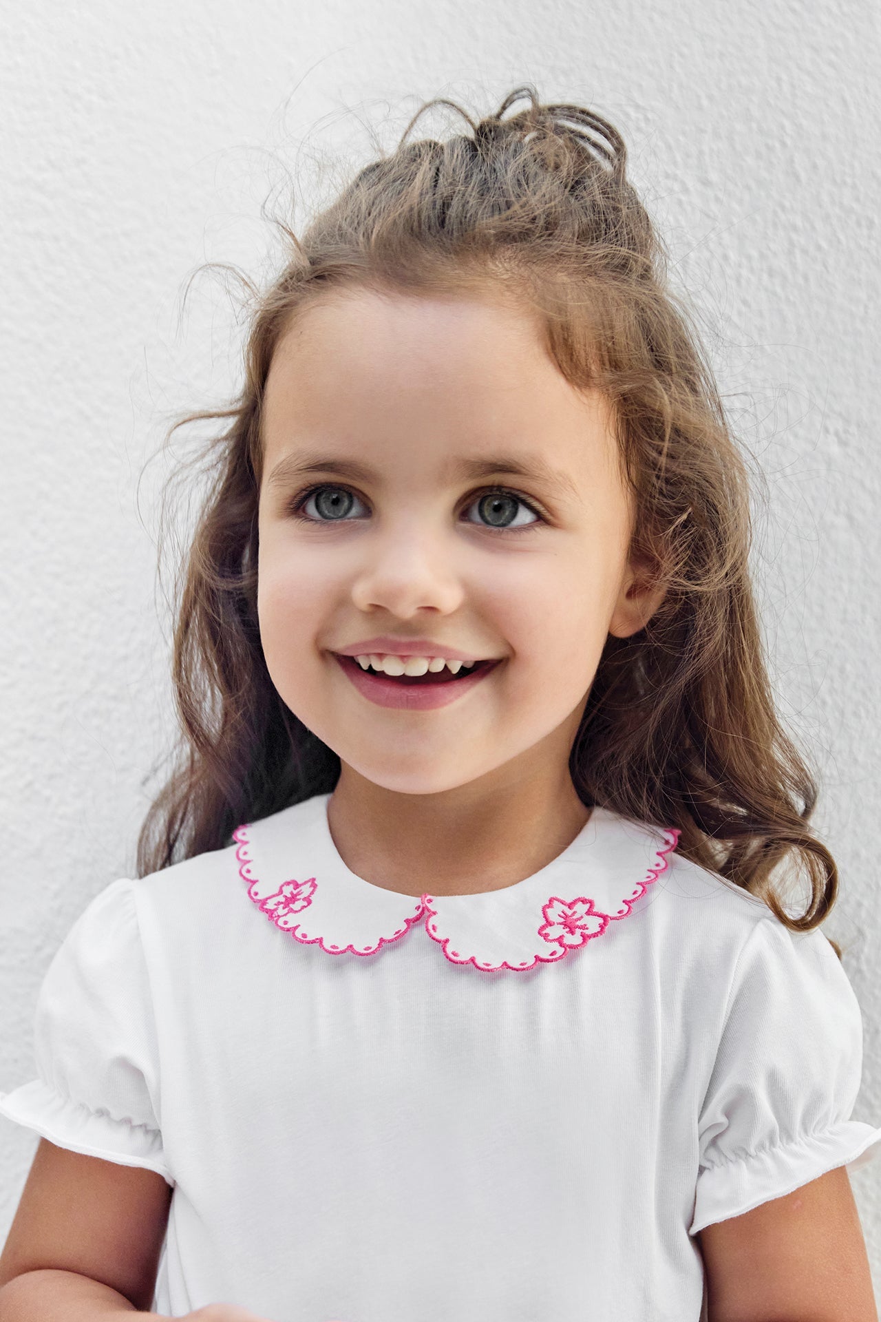 A young child with long brown hair and green eyes smiles while wearing the Tartine et Chocolat Baby Girls’ Peter Pan Collared Onesie, featuring a delicately embroidered white collar with pink flowers, standing against a plain light-colored wall.