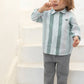 A young child stands barefoot on white stairs, wearing the Tartine et Chocolat Baby Boys’ and Toddler Badge Shirt—a soft cotton green and white stripe top—paired with gray pants, with one hand near their mouth, capturing an effortlessly casual look.