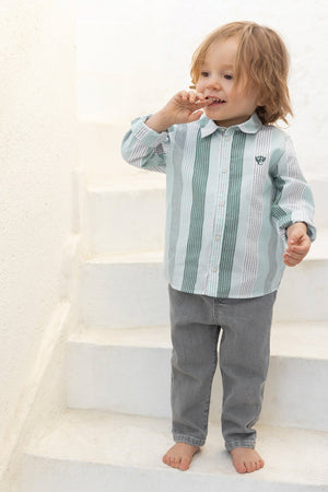 A young child stands barefoot on white stairs, wearing the Tartine et Chocolat Baby Boys’ and Toddler Badge Shirt—a soft cotton green and white stripe top—paired with gray pants, with one hand near their mouth, capturing an effortlessly casual look.