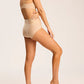 A woman models the Commando Zone Smoothing Brief in beige, showcasing its high-rise waist as she stands barefoot against a plain background with one leg slightly raised and her hand resting on her side.