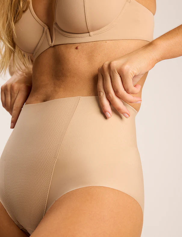 A woman models the Commando Zone Smoothing Brief in beige, a high-rise shapewear brief with smoothing panels, shown from shoulders to upper thighs against a plain background.