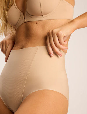A woman models the Commando Zone Smoothing Brief in beige, a high-rise shapewear brief with smoothing panels, shown from shoulders to upper thighs against a plain background.