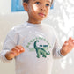A young child stands outdoors in soft sunlight wearing the Tartine et Chocolat Baby Boys’ and Boys’ Dinosaur Sweatshirt in grey heather, crafted from 100% cotton and adorned with a playful dinosaur illustration and text.