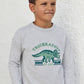 A young boy stands against a white textured wall, wearing the Tartine et Chocolat Boys’ Heather Sweatshirt, designed for kids with a cozy cotton feel and featuring a green dinosaur illustration with the words “Triceratops Dino Club.”.