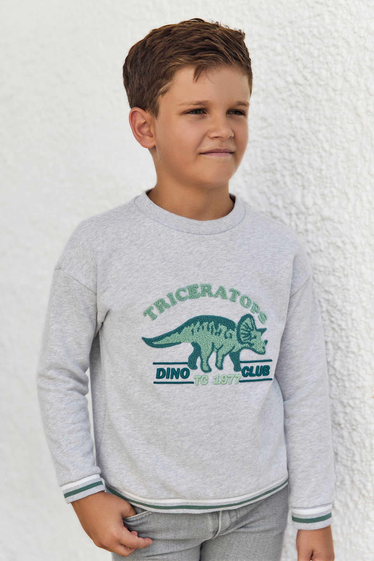 A young boy stands against a white textured wall, wearing the Tartine et Chocolat Boys’ Heather Sweatshirt, designed for kids with a cozy cotton feel and featuring a green dinosaur illustration with the words “Triceratops Dino Club.”.