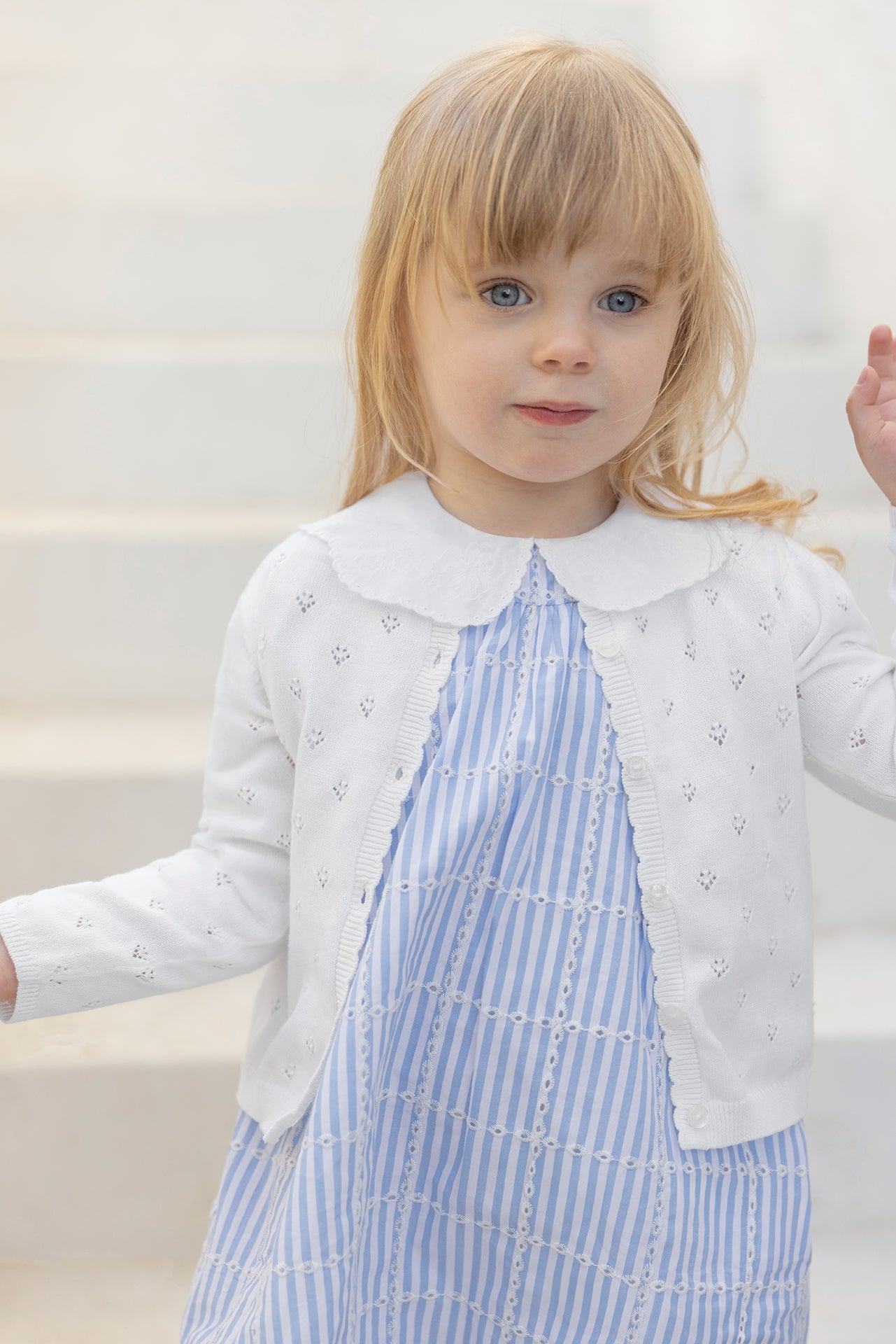 A young child with light brown hair stands in front of light-colored steps, wearing the Tartine et Chocolat Baby Girls’ and Girls’ Openwork Cardigan over a blue striped dress—showcasing the perfect lightweight layering piece for any occasion.