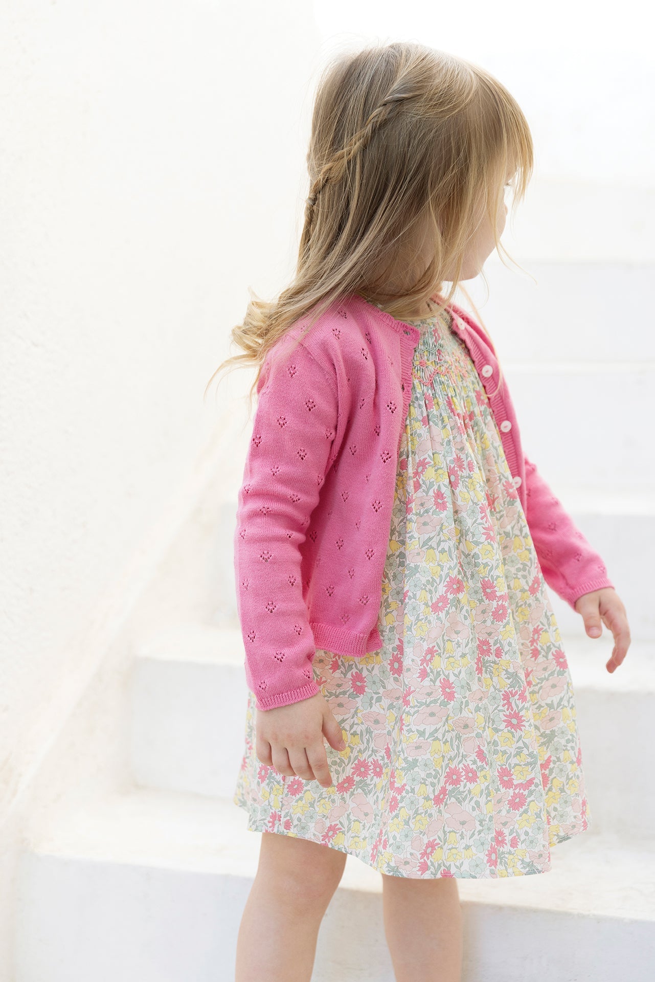 A young girl with light brown hair stands on white steps, looking to the side, wearing a pink Tartine et Chocolat Baby Girls’ and Girls’ Openwork Cardigan over a floral dress.