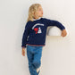 A young child with long blond hair wears a navy “Badminton” sweatshirt, Tartine et Chocolat Boys’ Cotton Jeans, and sneakers, leaning casually against a white wall with one arm resting on a protruding ledge.