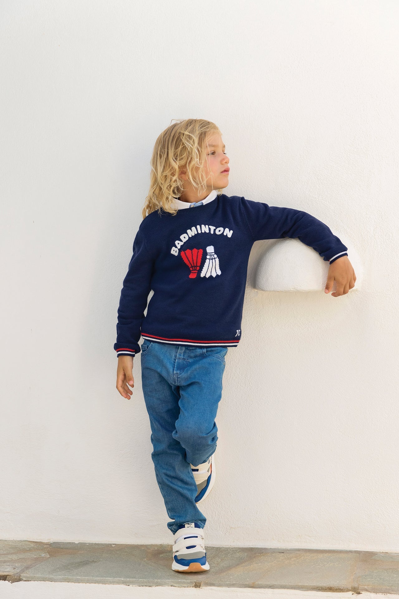 A young child with long blond hair wears a navy “Badminton” sweatshirt, Tartine et Chocolat Boys’ Cotton Jeans, and sneakers, leaning casually against a white wall with one arm resting on a protruding ledge.