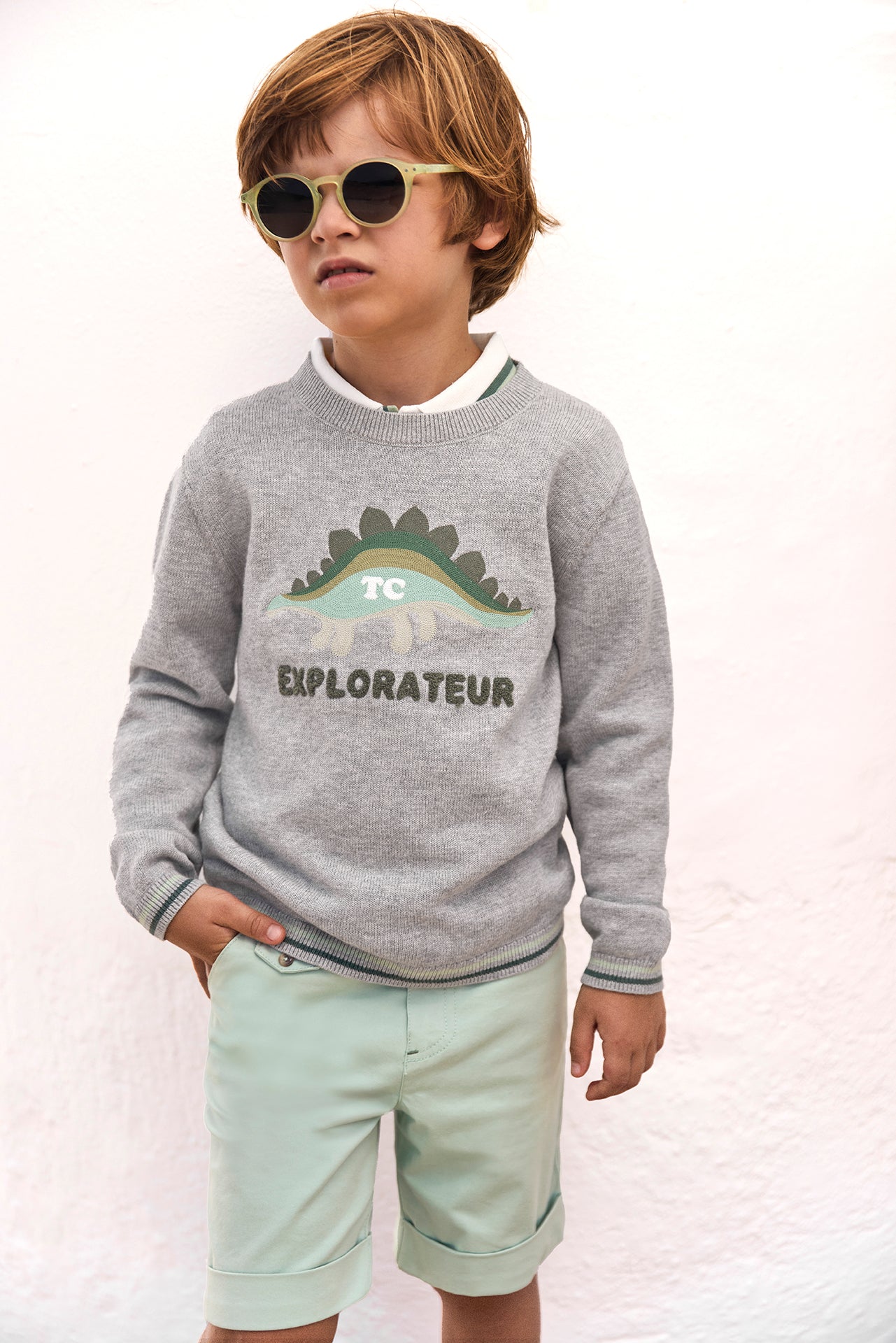 A young boy wearing sunglasses, the Tartine et Chocolat Boys’ Illustration Sweater made from 100% cotton featuring a dinosaur and the word “EXPLORATEUR,” and light green shorts stands against a plain white background.