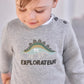 A toddler looks down while wearing the Tartine et Chocolat Baby Boys’ and Toddler Knitted Sweater, crafted from 100% cotton in grey with a dinosaur graphic and “EXPLORATEUR” printed on the front.