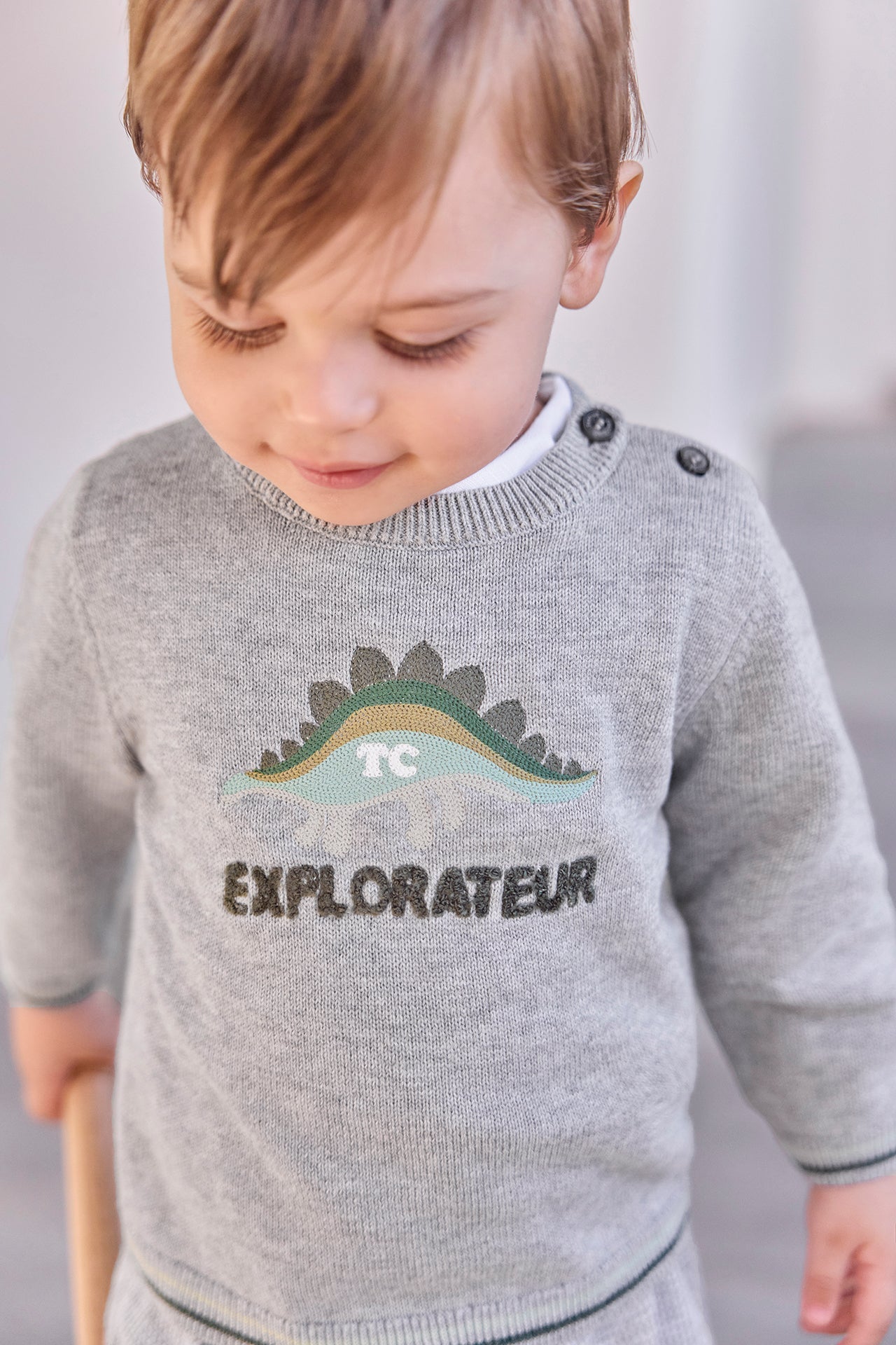 A toddler looks down while wearing the Tartine et Chocolat Baby Boys’ and Toddler Knitted Sweater, crafted from 100% cotton in grey with a dinosaur graphic and “EXPLORATEUR” printed on the front.