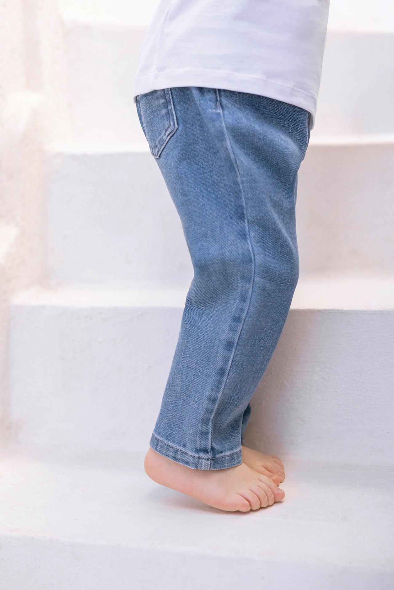 A little explorer wearing Tartine et Chocolat Baby Boys’ and Boys’ John Blue Jeans is walking up white stairs barefoot, paired with a crisp white shirt.