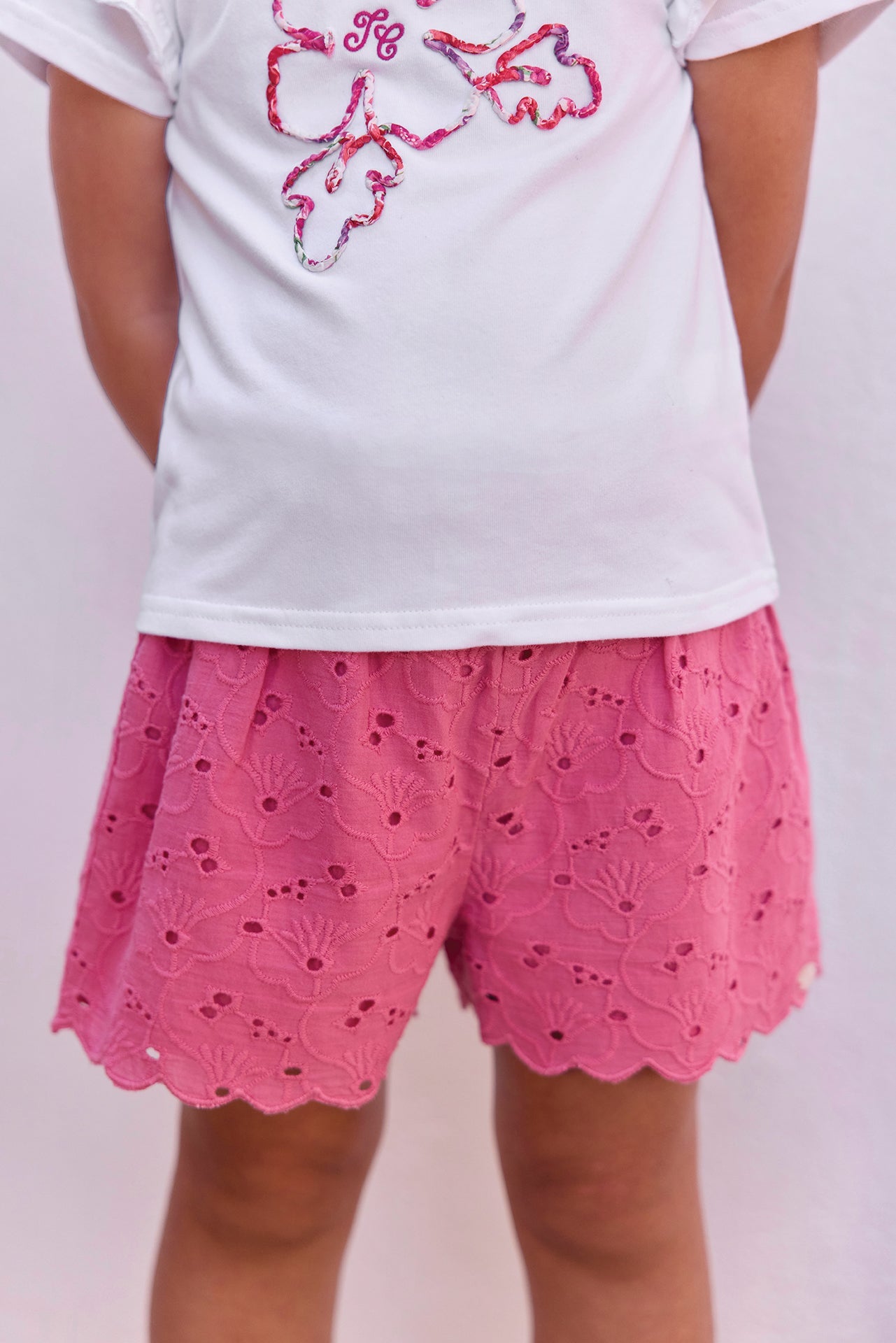 A child stands facing away against a plain light background, dressed in Tartine et Chocolat Baby Girls’ and Girls’ English Embroidery Shorts—white shorts featuring delicate embroidery and a scalloped hem—paired with a simple white T-shirt.