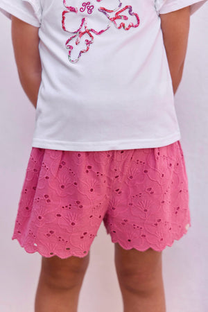A child stands facing away against a plain light background, dressed in Tartine et Chocolat Baby Girls’ and Girls’ English Embroidery Shorts—white shorts featuring delicate embroidery and a scalloped hem—paired with a simple white T-shirt.