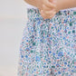 A child wearing Tartine et Chocolat’s Baby Girls’ and Girls’ Liberty Short, featuring a delicate floral print and a crisp white shirt, stands with their hands clasped together in front of them.