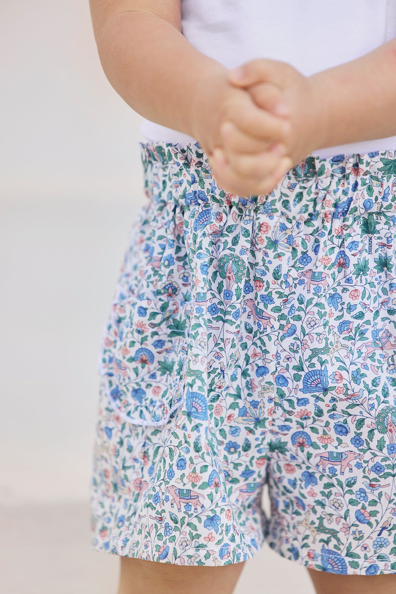 A child wearing Tartine et Chocolat’s Baby Girls’ and Girls’ Liberty Short, featuring a delicate floral print and a crisp white shirt, stands with their hands clasped together in front of them.