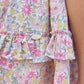 Close-up of a ruffled Girls’ Liberty Fabric Print Skirt by Tartine et Chocolat, featuring a garden-inspired floral pattern in shades of pink, yellow, blue, and white, with a small white button detail. Skin is visible on the right side of the image.