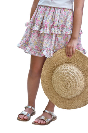 A child wearing the Tartine et Chocolat Girls’ Liberty Fabric Print Skirt and silver sandals holds a large straw hat, standing against a white background.