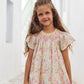 A young girl with long brown hair stands in a narrow, sunlit alley with white walls and pastel green stairs in the background, wearing the Tartine et Chocolat Girls’ Liberty Print Dress featuring a smocked collar.