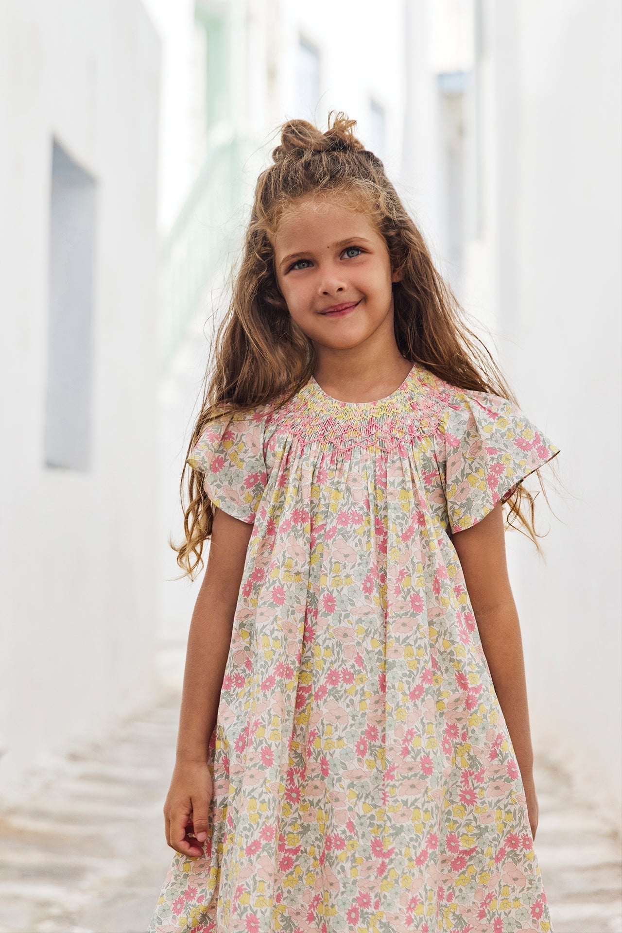 A young girl with long brown hair stands in a narrow, sunlit alley with white walls and pastel green stairs in the background, wearing the Tartine et Chocolat Girls’ Liberty Print Dress featuring a smocked collar.