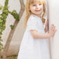 A young girl with blonde hair is standing outside by a white wall, smiling at the camera, wearing the Tartine et Chocolat Baby Girls’ and Girls’ English Embroidery Cotton Dress.