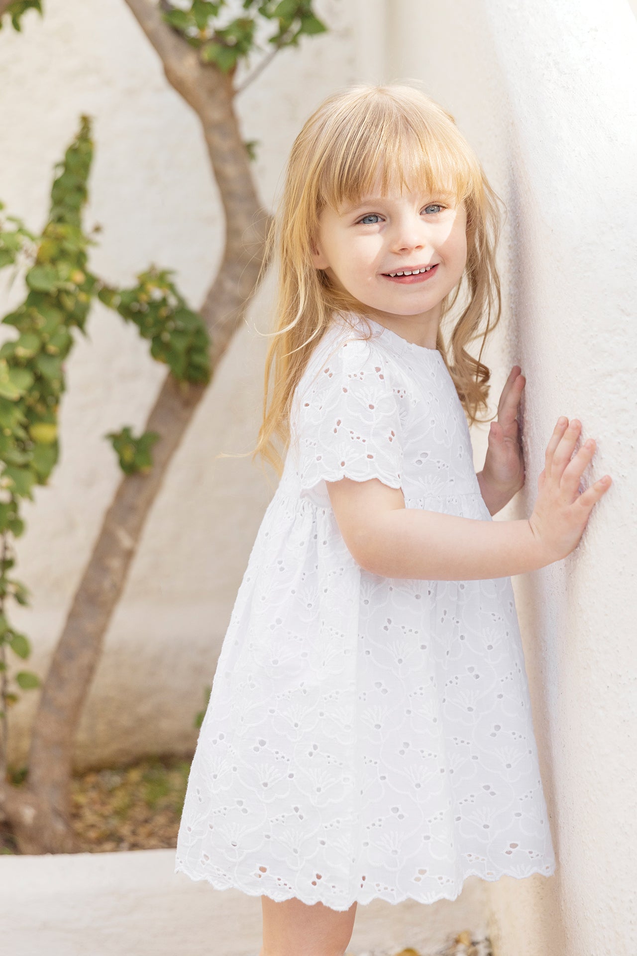 A young girl with blonde hair is standing outside by a white wall, smiling at the camera, wearing the Tartine et Chocolat Baby Girls’ and Girls’ English Embroidery Cotton Dress.