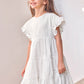 A young girl wears the Tartine et Chocolat Girls’ Openwork Dress with ruffled sleeves, standing and smiling by a white wall adorned with pink flowers.