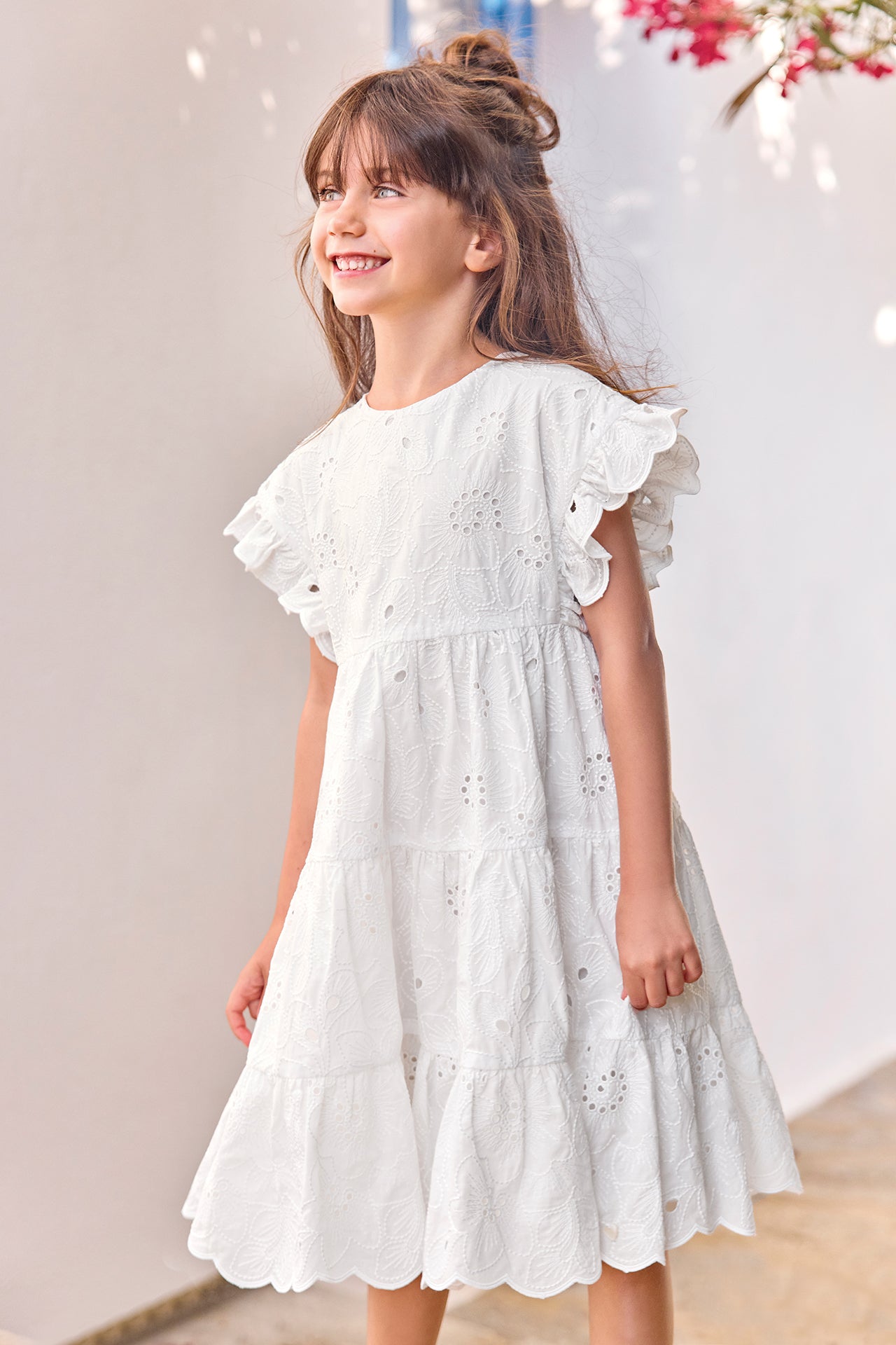 A young girl wears the Tartine et Chocolat Girls’ Openwork Dress with ruffled sleeves, standing and smiling by a white wall adorned with pink flowers.
