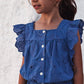 A young girl with brown hair wears the Tartine et Chocolat Girls’ Openwork Short Jumpsuit, featuring short sleeves, button-up front, delicate eyelet detailing, and ruffles, as she stands against a textured white wall.