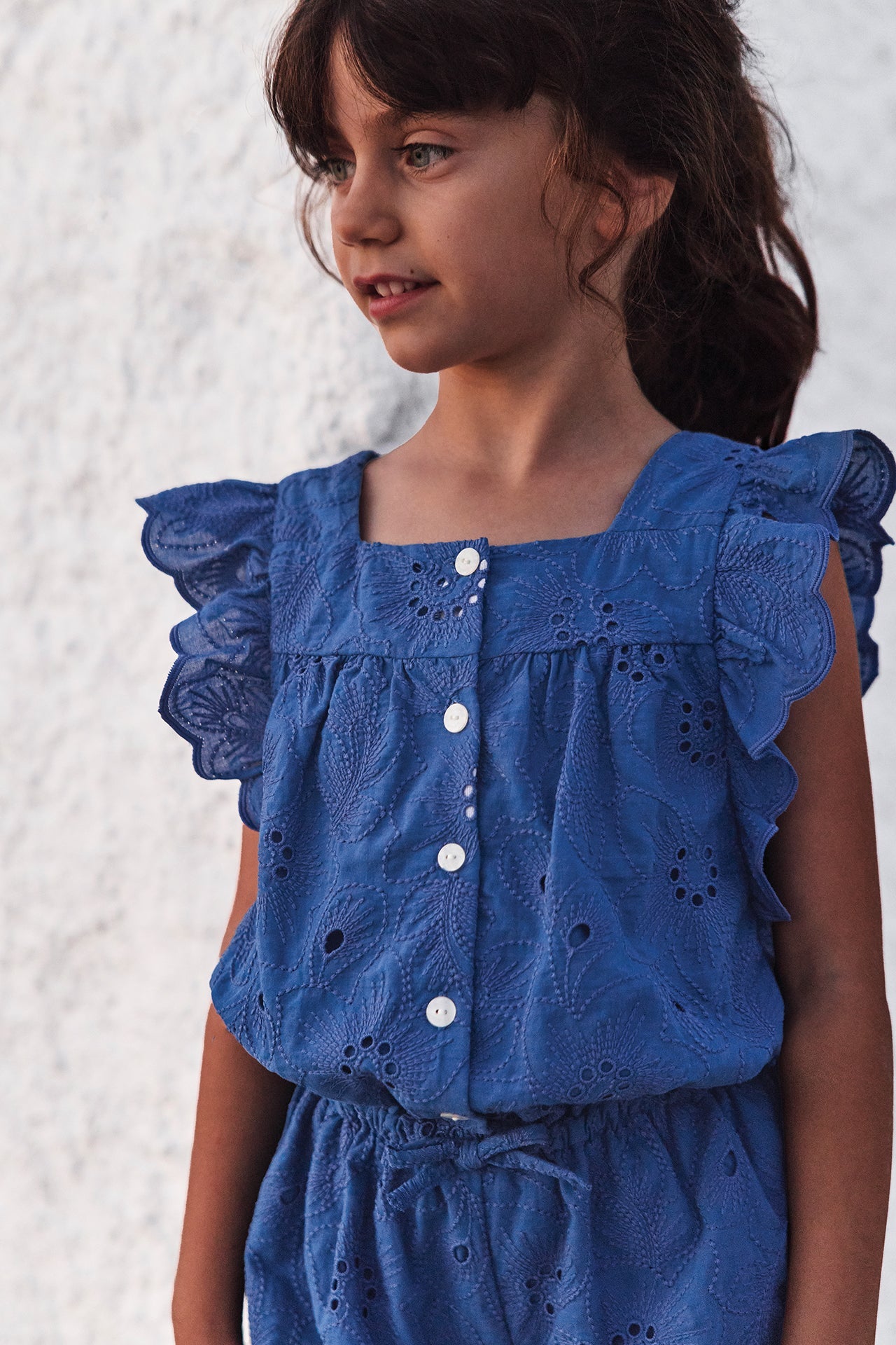 A young girl with brown hair wears the Tartine et Chocolat Girls’ Openwork Short Jumpsuit, featuring short sleeves, button-up front, delicate eyelet detailing, and ruffles, as she stands against a textured white wall.