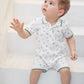A young child with light brown hair sits barefoot on white steps, dressed in the Tartine et Chocolat Baby Boys’ Short Jumpsuit—a cozy cotton set featuring a white short-sleeve polo shirt and matching shorts adorned with a small colorful print.