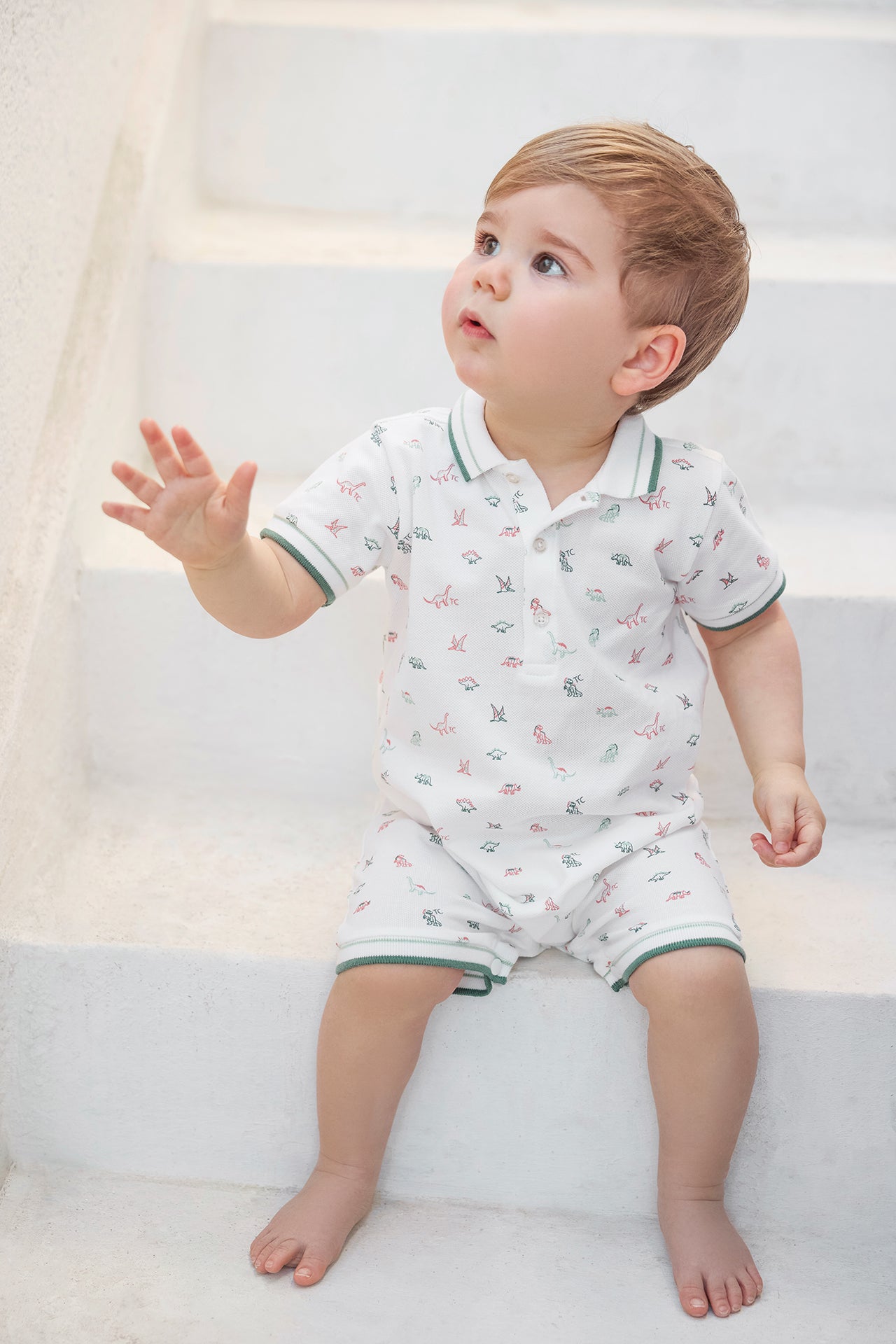 A young child with light brown hair sits barefoot on white steps, dressed in the Tartine et Chocolat Baby Boys’ Short Jumpsuit—a cozy cotton set featuring a white short-sleeve polo shirt and matching shorts adorned with a small colorful print.