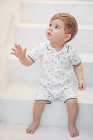 A young child with light brown hair sits barefoot on white steps, dressed in the Tartine et Chocolat Baby Boys’ Short Jumpsuit—a cozy cotton set featuring a white short-sleeve polo shirt and matching shorts adorned with a small colorful print.