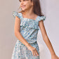 A young girl with long brown hair stands smiling against a light background, pink flowers visible in the upper right corner, wearing the Tartine et Chocolat Girls’ Liberty Print Short Jumpsuit with ruffled details and her right hand extended.