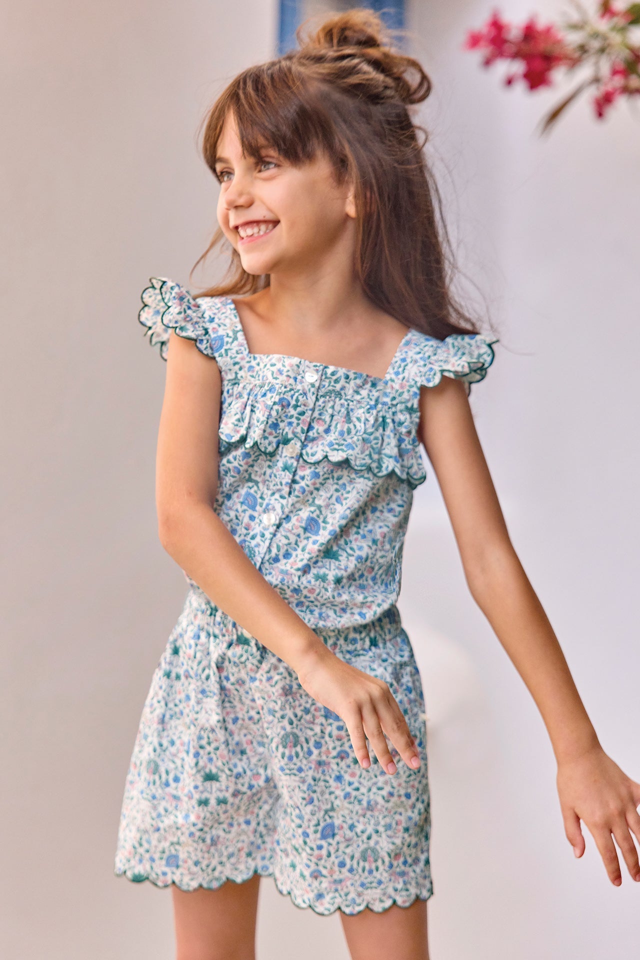 A young girl with long brown hair stands smiling against a light background, pink flowers visible in the upper right corner, wearing the Tartine et Chocolat Girls’ Liberty Print Short Jumpsuit with ruffled details and her right hand extended.