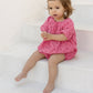 A toddler wearing the Tartine et Chocolat Baby Girls’ English Embroidery Bubble sits barefoot on white steps, hands clasped together, with a few red flower petals scattered on the step below.