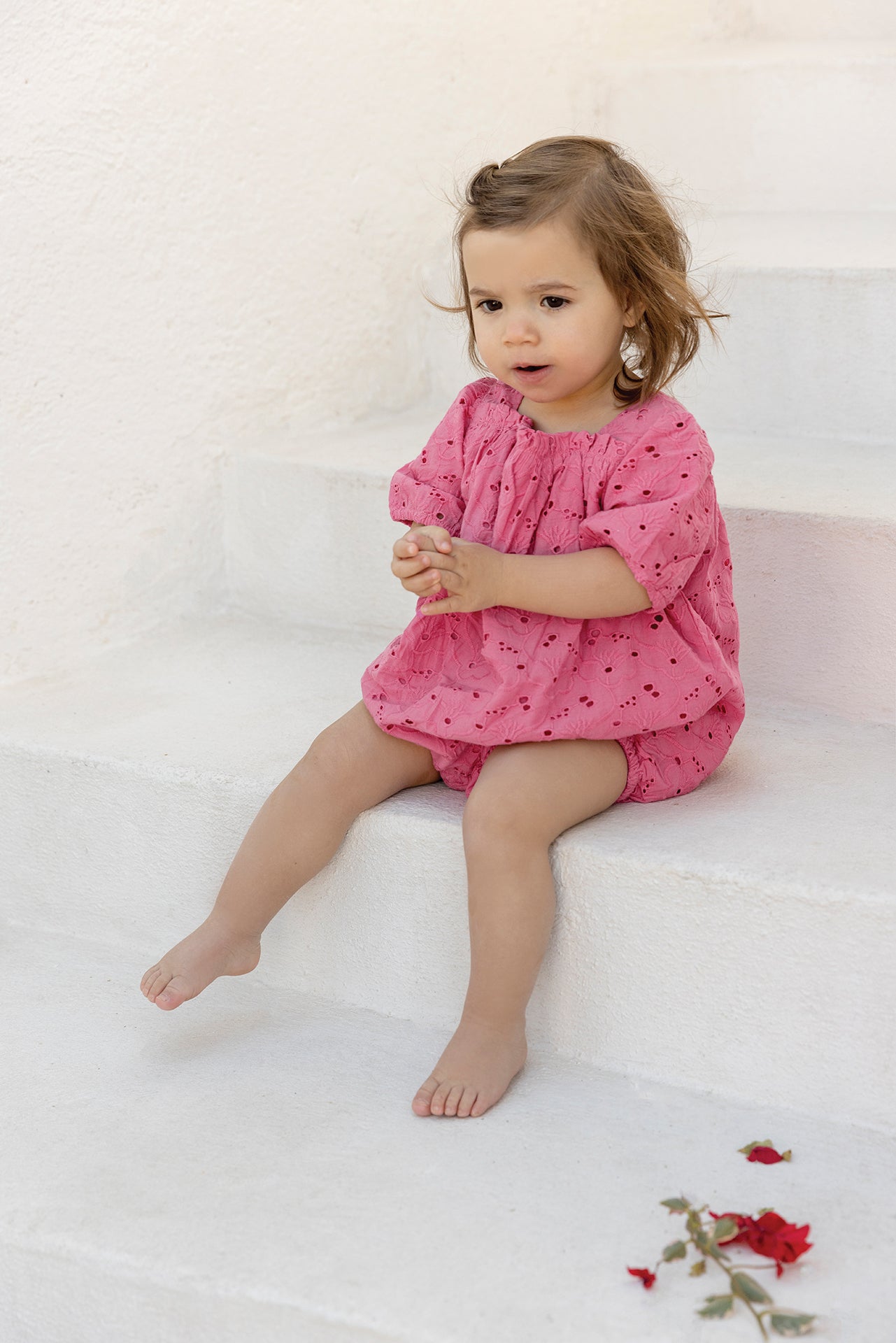 A toddler wearing the Tartine et Chocolat Baby Girls’ English Embroidery Bubble sits barefoot on white steps, hands clasped together, with a few red flower petals scattered on the step below.