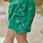 A child is standing outdoors wearing Tartine et Chocolat Baby Boys’ and Boys’ Swim Shorts in green, decorated with colorful dinosaur patterns. The shorts feature an elasticated waistband and a visible side pocket.