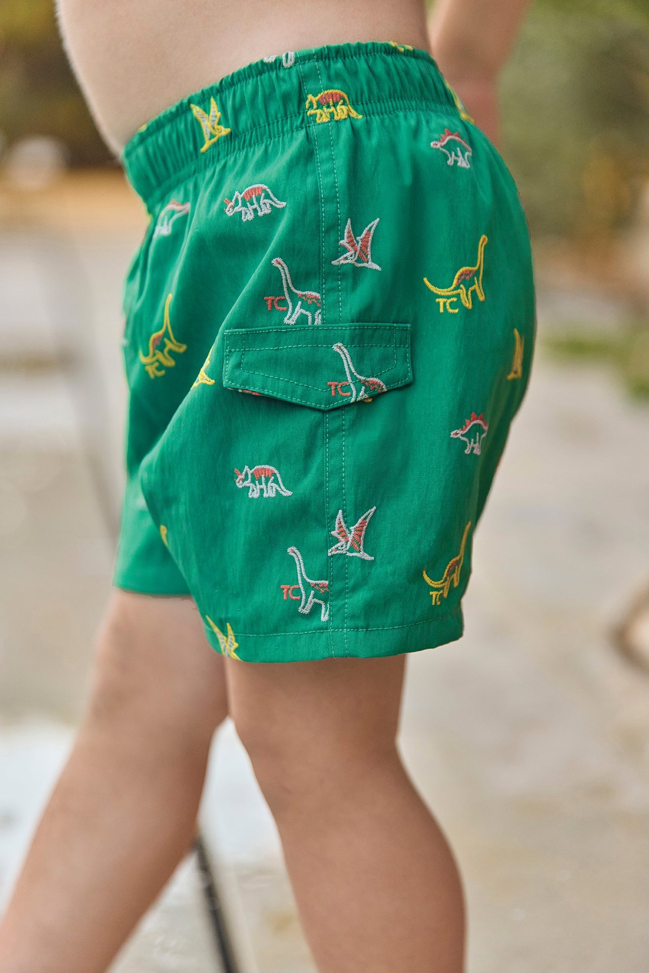 A child is standing outdoors wearing Tartine et Chocolat Baby Boys’ and Boys’ Swim Shorts in green, decorated with colorful dinosaur patterns. The shorts feature an elasticated waistband and a visible side pocket.