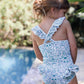 A young girl stands by a pool surrounded by greenery, her hair in a loose bun, wearing the Tartine et Chocolat Baby Girls’ and Girls’ Liberty Printed Swimsuit.
