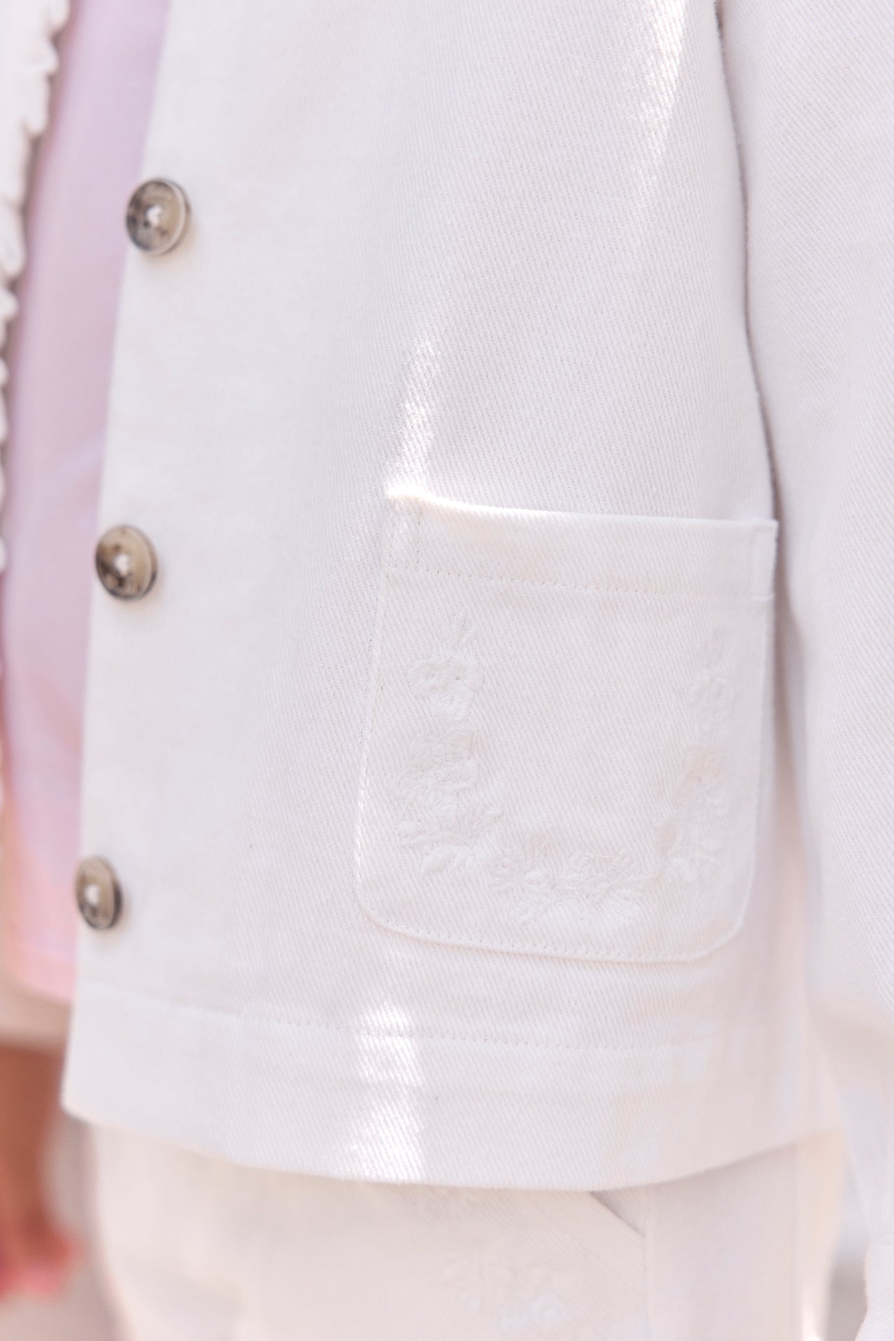 Close-up of the Tartine et Chocolat Girls’ Cotton Jacket in white, featuring floral embroidery on the pocket, ruffled edging, and brown buttons—shown as part of a matching outfit.