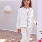 A young girl stands outdoors on a stone path near a white building with a blue window, wearing the Tartine et Chocolat Girls’ Cotton Jacket with embroidered pockets and matching pants.