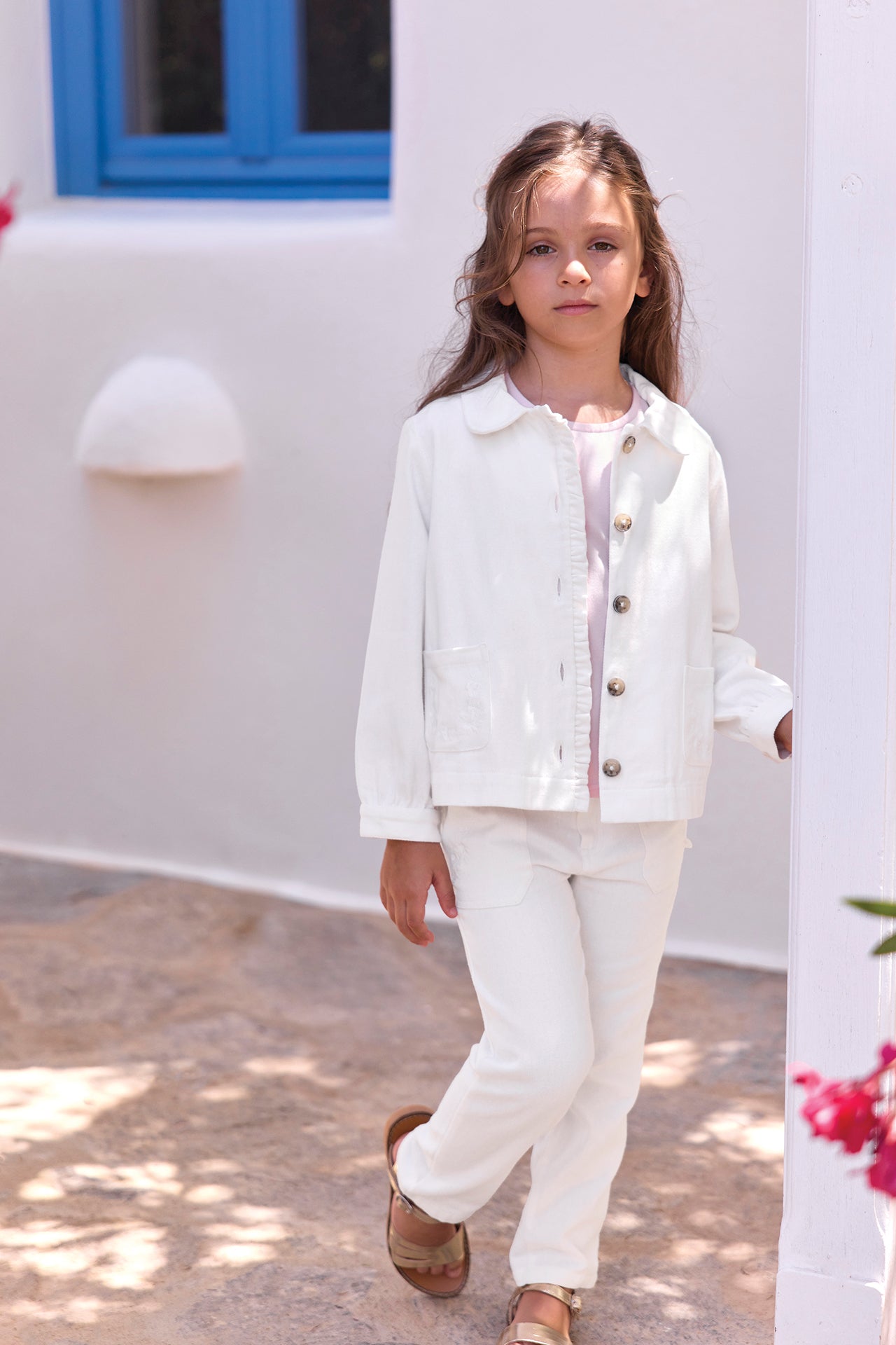 A young girl stands outdoors on a stone path near a white building with a blue window, wearing the Tartine et Chocolat Girls’ Cotton Jacket with embroidered pockets and matching pants.